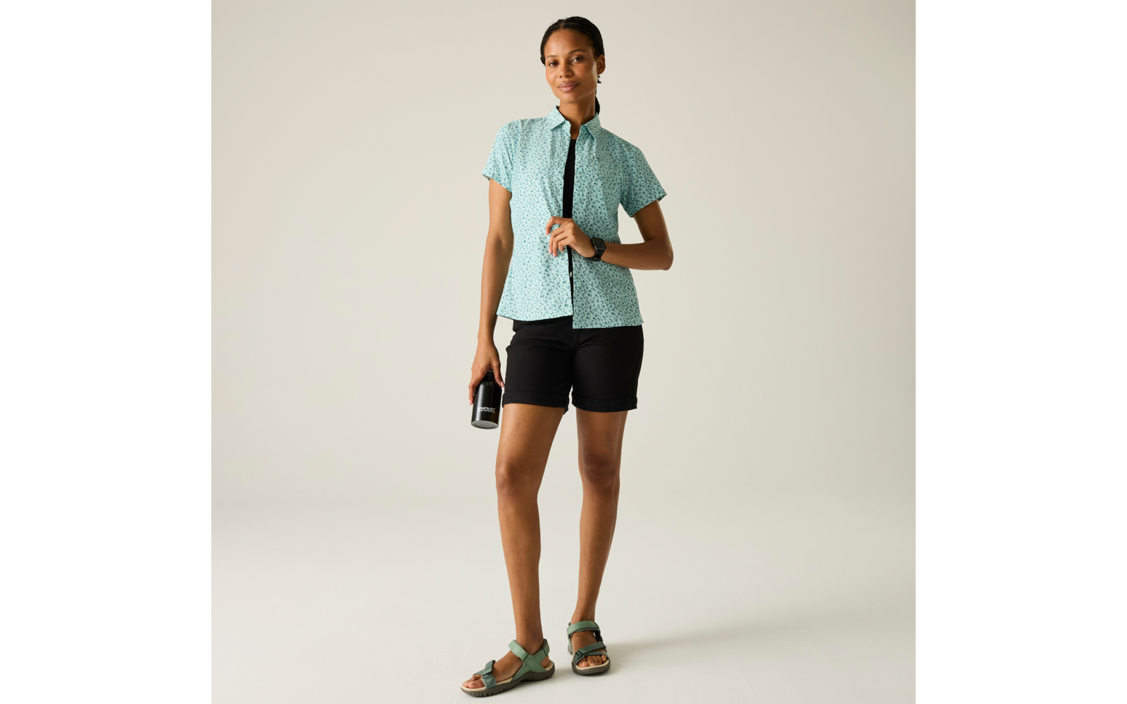 Person steht vor hellem Hintergrund, trägt mintgrünes kurzärmeliges gemustertes Hemd, schwarze Shorts, grüne Sandalen, hält schwarze Trinkflasche in rechter Hand, linke Hand am Hemd, trägt Armbanduhr und Ohrstecker, Haare zurückgebunden