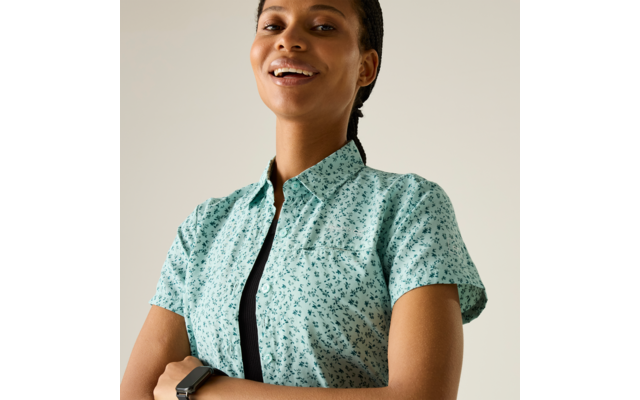 Person mit kurzärmligem, hellgrünem, gemustertem Hemd über einem schwarzen Top, Arme verschränkt, lächelnd vor hellem Hintergrund