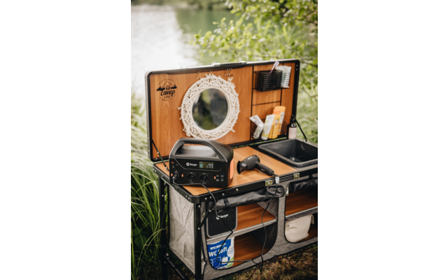 Malle ouverte à intérieur en bois, miroir rond blanc, unité d'alimentation portable avec écran numérique, sèche-cheveux, vasque, flacons de toilette, seau en plastique marqué WC, rive avec eau et plantes en arrière-plan