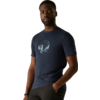 Persoon staat voor een beige achtergrond, draagt een donkerblauw T‑shirt met een lichte cirkelvormige print op de borst, één hand in de zak, kijkt naar de camera