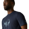 Close-up van het bovenlichaam van een man met een donkerblauw T-shirt met een witte illustratie van bergen en een wandelaar op de borst, beige achtergrond
