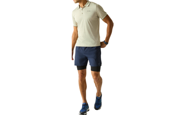 Mann in hellgrünem Poloshirt, dunkelblauen Shorts mit schwarzer Innenlage, blauen Laufschuhen und Armbanduhr, Blick nach links vor hellem Hintergrund