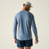 Person von hinten, trägt hellblaues Langarmshirt, dunkelblaue Shorts und weiße Kappe mit Sonnenbrille; heller neutraler Hintergrund