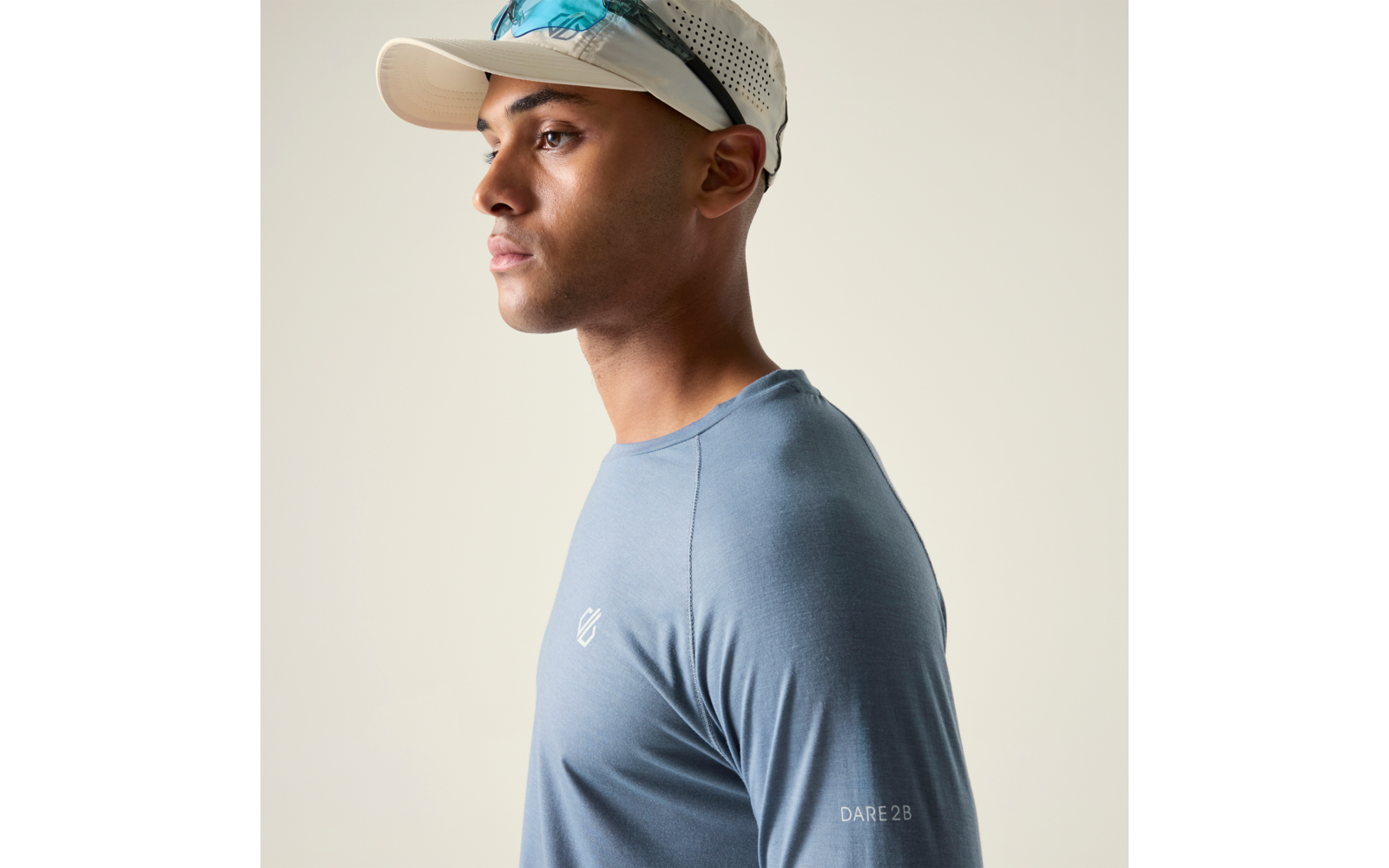 Seitliches Porträt einer Person mit blauem Sportshirt, weißer Kappe und Sonnenbrille auf der Kappe vor hellem Hintergrund