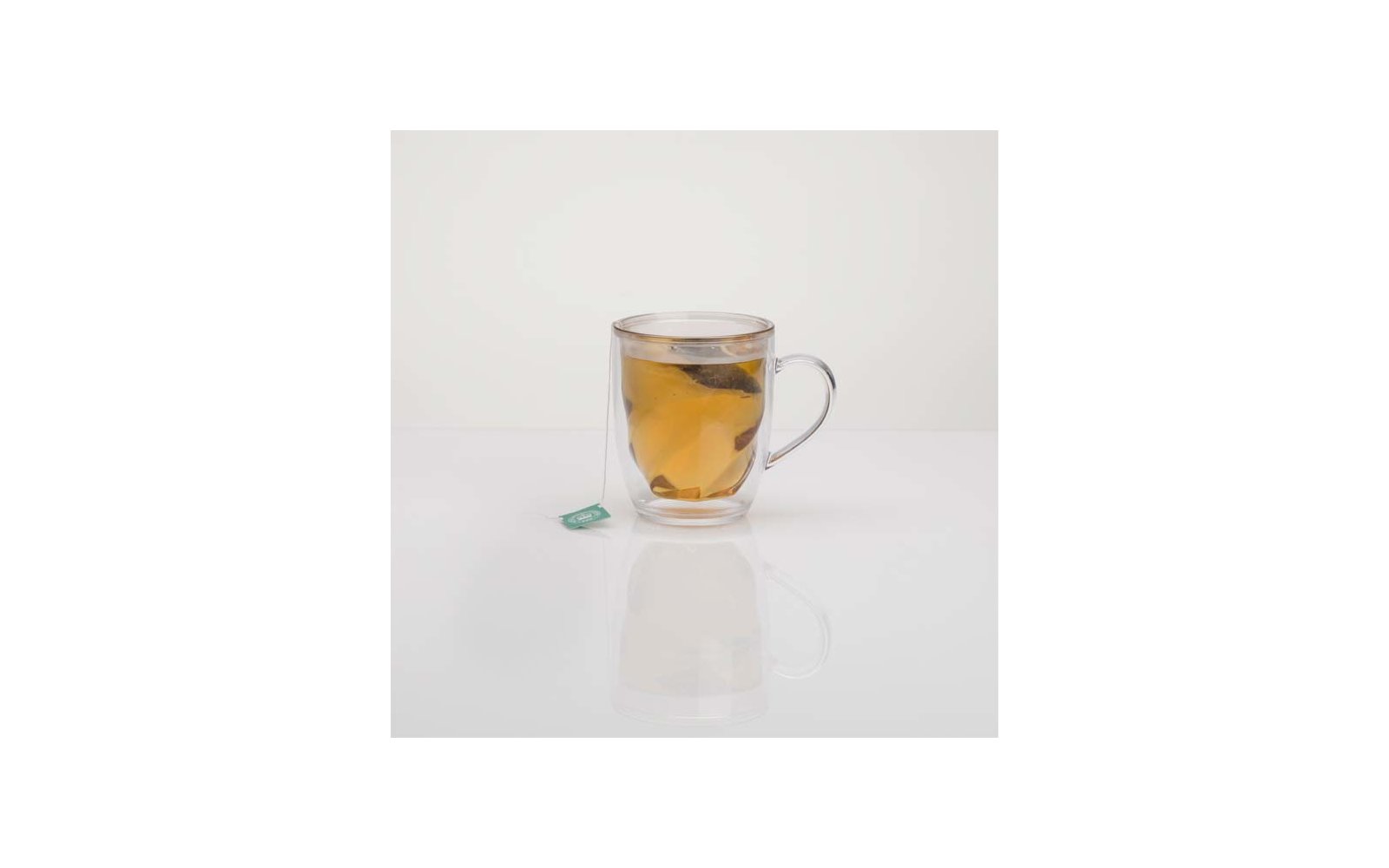 Tasse en verre double paroi avec sachet de thé et liquide ambré sur une surface blanche, étiquette verte du sachet à gauche, reflet visible
