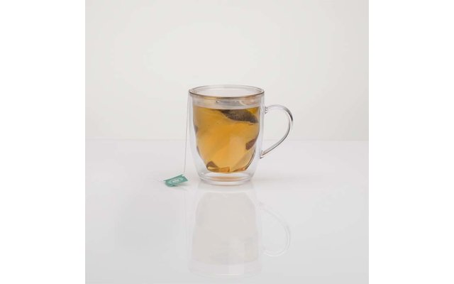 Tasse en verre double paroi avec sachet de thé et liquide ambré sur une surface blanche, étiquette verte du sachet à gauche, reflet visible