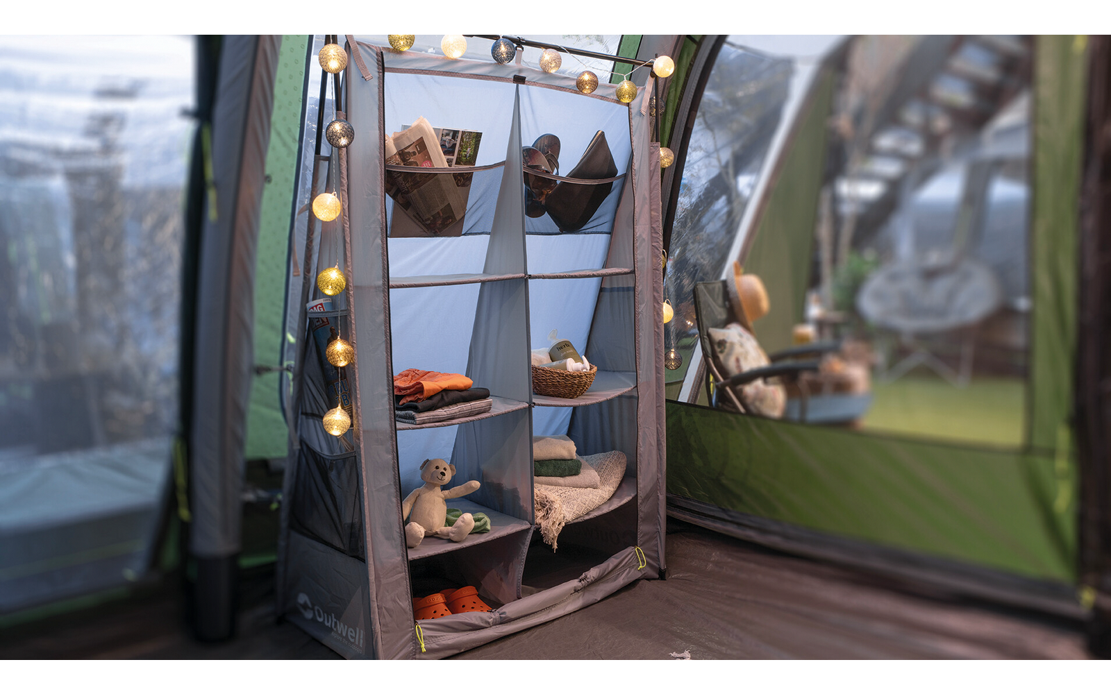 Regal mit acht Fächern in einem Zelt, darin Stoffkörbe, gefaltete Kleidung, Schuhe und ein Teddybär; Lichterkette an der linken Regalseite; Zeltwände und ein hängender Sitz im Hintergrund