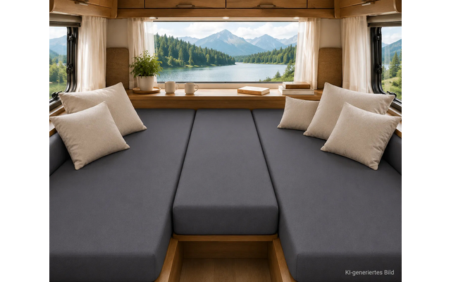 Interieur van een camper met een donkergrijze lig-/zitplaats en meerdere beige kussens; vensterbank met een potplant, twee kopjes en een boek; groot raam toont een meer, beboste heuvels en bergtoppen