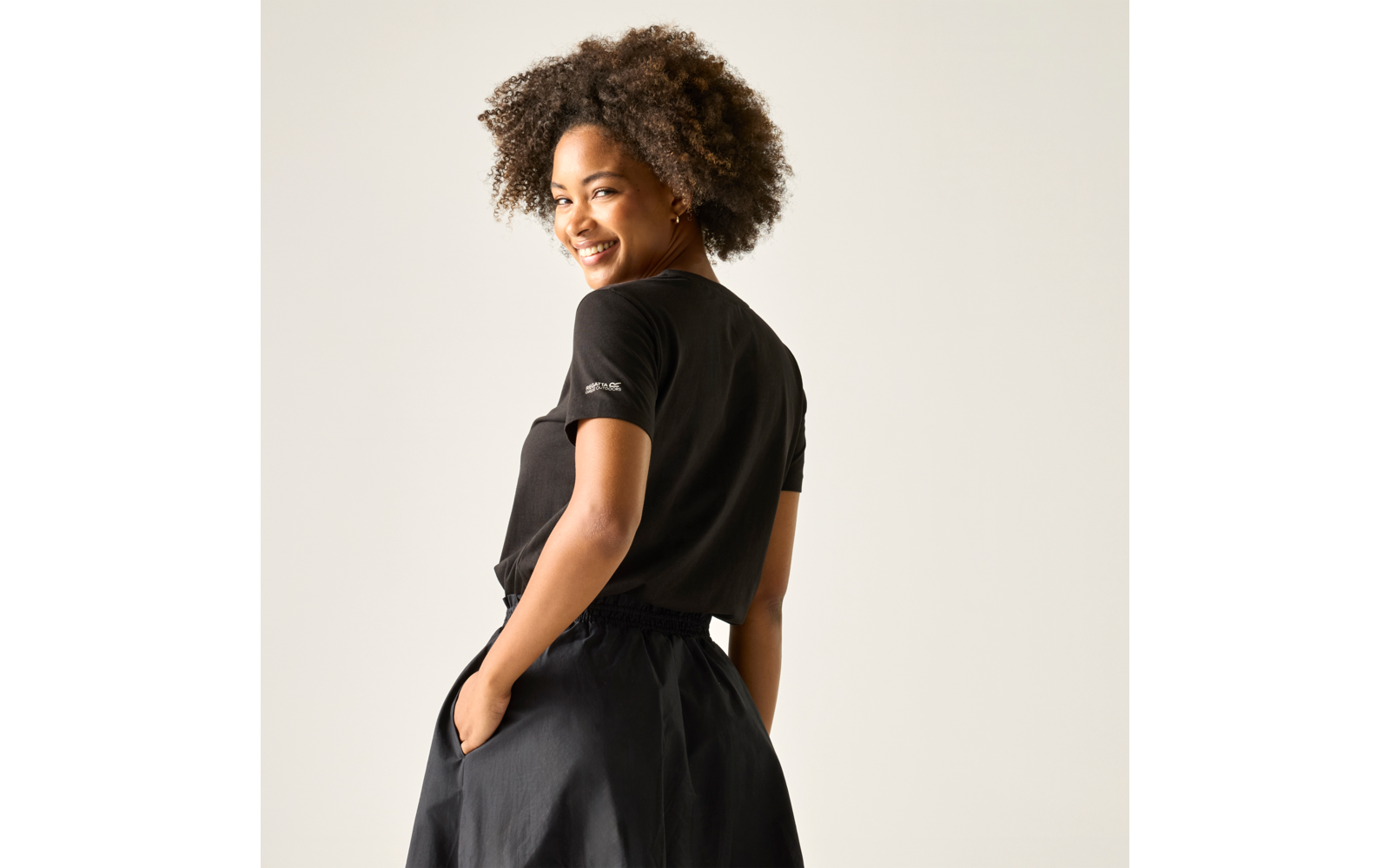 Person mit vollem, lockigem Haar trägt schwarzes T‑Shirt und schwarze Rockbekleidung, steht mit Händen in den Taschen und schaut über die Schulter vor einem hellen Hintergrund