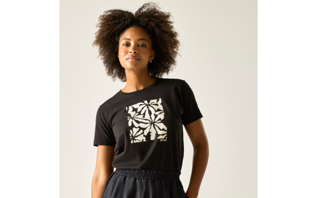 Person mit voluminösen, lockigen Haaren trägt schwarzes T‑Shirt mit weißem, floralem Quadratmotiv und dunkle Hose, steht vor hellem Hintergrund, schaut zur Kamera