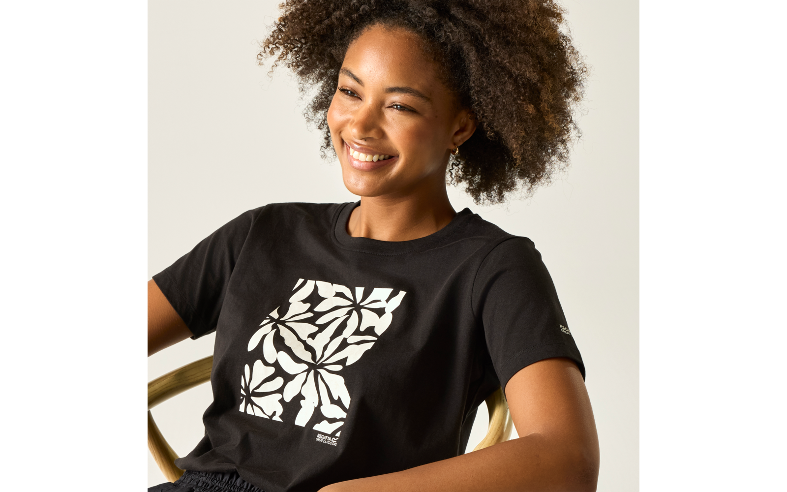 Lächelnde Person mit lockigem Haar, trägt ein schwarzes T‑Shirt mit weißem floralen Quadratdruck, sitzt auf einem Stuhl mit goldener Armlehne vor hellem Hintergrund