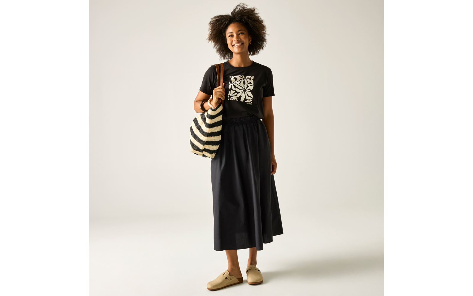 Frau steht, lächelt, schwarzes T-Shirt mit weißem Muster, schwarzer Midirock, große schwarz-weiß gestreifte Tasche über der Schulter, beige Schuhe, heller Hintergrund