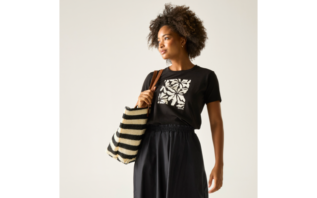 Person mit kurzem, lockigem Afrohaar, schwarzem T‑Shirt mit weißem Blumenquadratmuster, schwarzem Rock, beige‑schwarz gestreifter Tasche über der Schulter, heller Hintergrund, Blick zur Seite