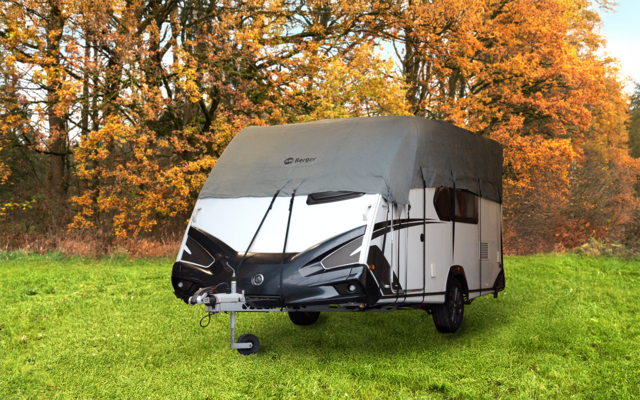 Caravan with gray cover on green grass in front of autumn trees