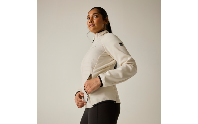 Persona con giacca beige chiaro a cuciture orizzontali e leggings scuri, in vista tre quarti, mani sulla cerniera, capelli raccolti in coda, sfondo chiaro uniforme