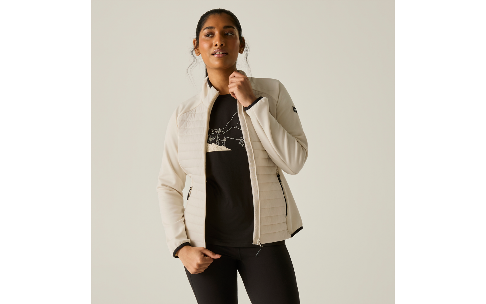 Persona davanti a uno sfondo chiaro, indossa una giacca trapuntata beige su una t‑shirt nera con motivo montagna, leggings neri, tiene la cerniera della giacca con una mano