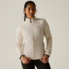 Persona con capelli raccolti indossa una giacca con zip trapuntata chiara con logo visibile e leggings neri, in piedi su sfondo uniforme, mani sull'orlo