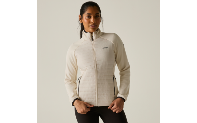 Persona con capelli raccolti indossa una giacca con zip trapuntata chiara con logo visibile e leggings neri, in piedi su sfondo uniforme, mani sull'orlo