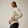 Persona con giacca beige chiaro a cuciture orizzontali e leggings scuri, in vista tre quarti, mani sulla cerniera, capelli raccolti in coda, sfondo chiaro uniforme