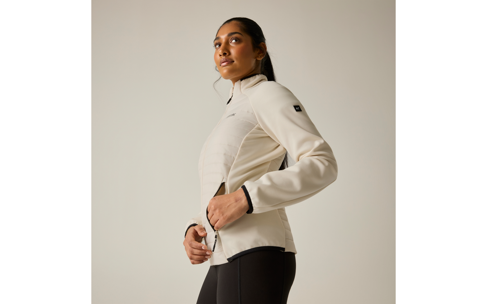 Persona con giacca beige chiaro a cuciture orizzontali e leggings scuri, in vista tre quarti, mani sulla cerniera, capelli raccolti in coda, sfondo chiaro uniforme