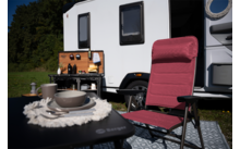 Roter Campingstuhl vor weißem Wohnmobil mit geöffneter Außenküche; daneben Tisch mit Teller, Schüssel und Tasse, Gras und Bäume im Hintergrund