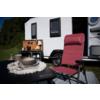 Rode campingstoel voor een witte camper met open buitenkeuken; naast de stoel een tafel met bord, kom en kopje, gras en bomen op de achtergrond