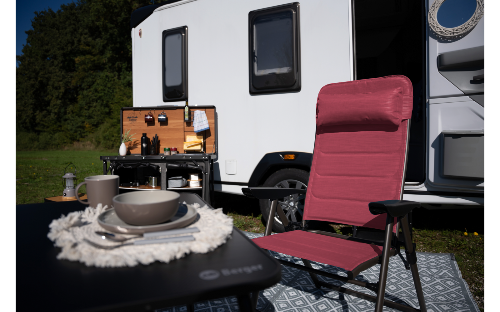 Rode campingstoel voor een witte camper met open buitenkeuken; naast de stoel een tafel met bord, kom en kopje, gras en bomen op de achtergrond