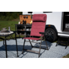 Rode opvouwbare campingstoel met hoofdsteun op een patroonmat voor een witte camper, naast een tafel met servies en een open buitenkeuken met pannen en flessen