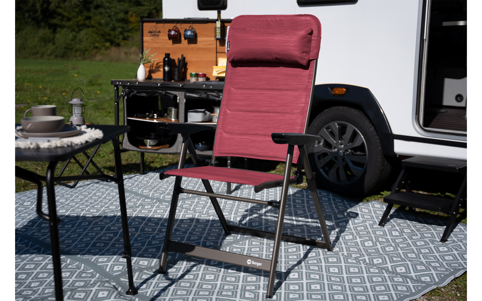 Rode opvouwbare campingstoel met hoofdsteun op een patroonmat voor een witte camper, naast een tafel met servies en een open buitenkeuken met pannen en flessen