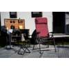 Fauteuil de camping pliant bordeaux avec accoudoirs sur un tapis à motifs devant une caravane ; à gauche une petite table pliante avec une bouilloire argentée, un réchaud et des ustensiles de cuisson, au-dessus une kitchenette en bois ouverte avec bouteilles et ustensiles ; caravane avec deux fenêtres et porte ouverte