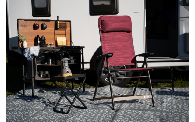 Fauteuil de camping pliant bordeaux avec accoudoirs sur un tapis à motifs devant une caravane ; à gauche une petite table pliante avec une bouilloire argentée, un réchaud et des ustensiles de cuisson, au-dessus une kitchenette en bois ouverte avec bouteilles et ustensiles ; caravane avec deux fenêtres et porte ouverte