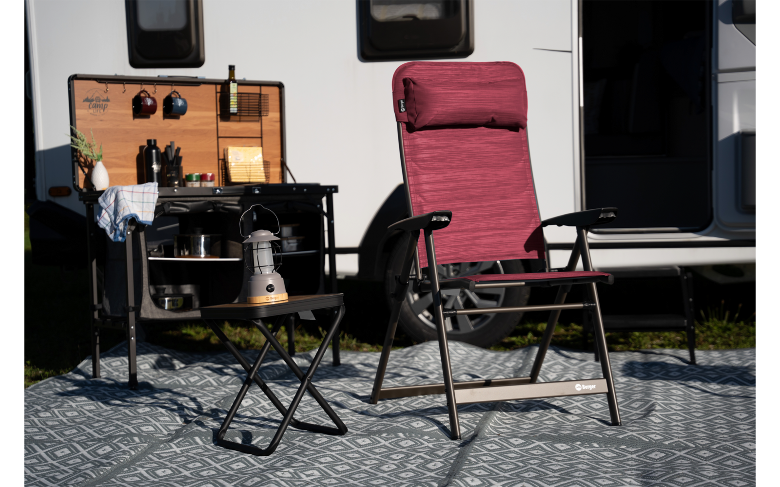 Fauteuil de camping pliant bordeaux avec accoudoirs sur un tapis à motifs devant une caravane ; à gauche une petite table pliante avec une bouilloire argentée, un réchaud et des ustensiles de cuisson, au-dessus une kitchenette en bois ouverte avec bouteilles et ustensiles ; caravane avec deux fenêtres et porte ouverte