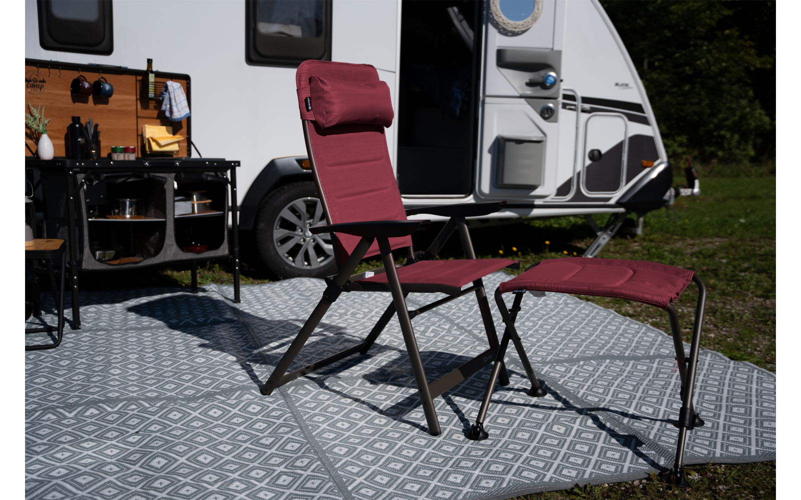 Rode vouwstoel met bijpassende voetensteun op patroonmat voor een geparkeerde caravan met open deur en plank met kookgerei