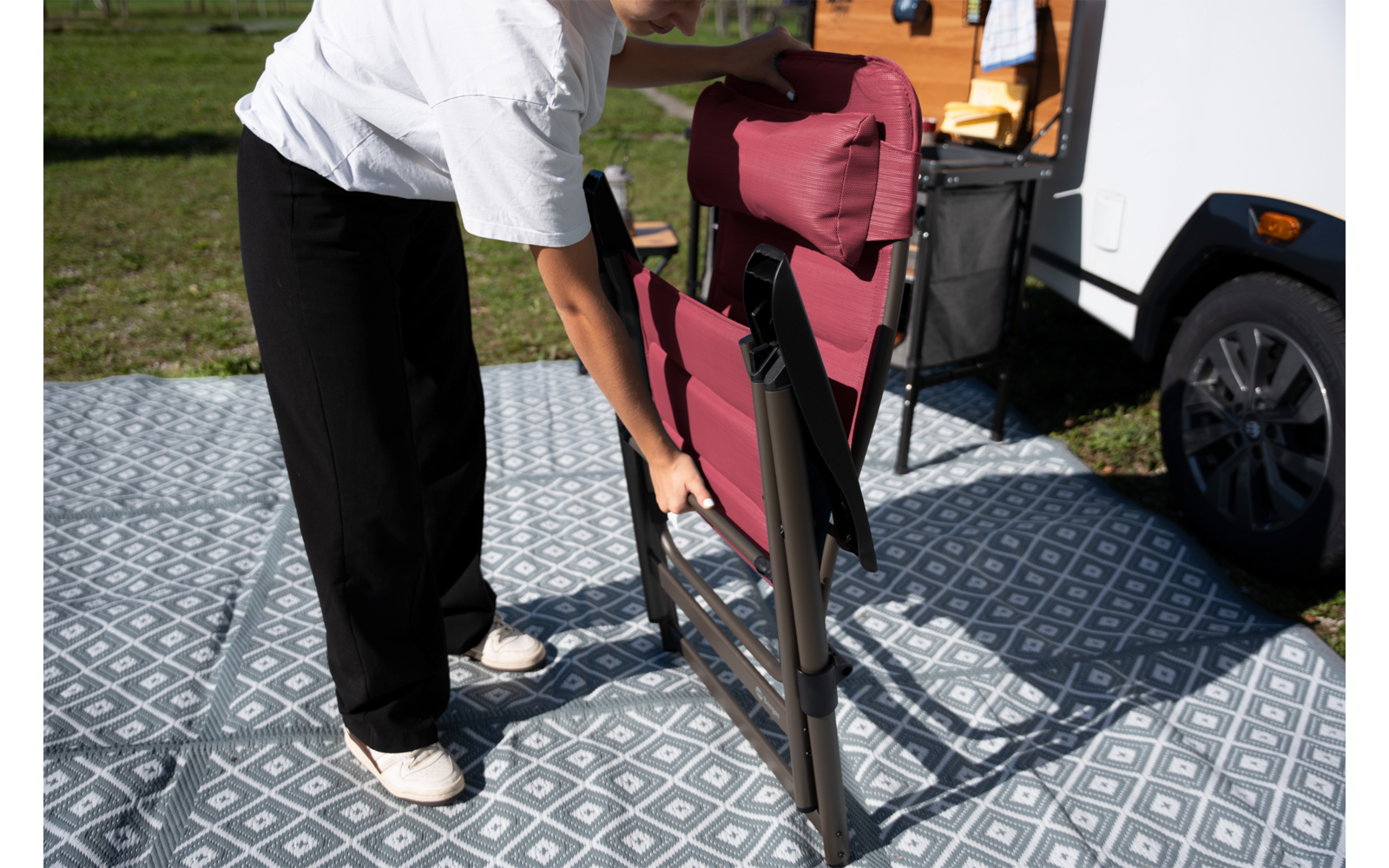 Personne tenant une chaise pliante rouge à côté d’un camping-car blanc sur un tapis extérieur gris à motifs