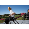Personne aux cheveux blonds attachés, en t‑shirt blanc et pantalon noir, assise sur une chaise pliante rouge et regardant vers la gauche. Tapis à motifs au premier plan, champ vert et ciel bleu en arrière‑plan, deuxième chaise et matériel de camping près d’un bâtiment en bois