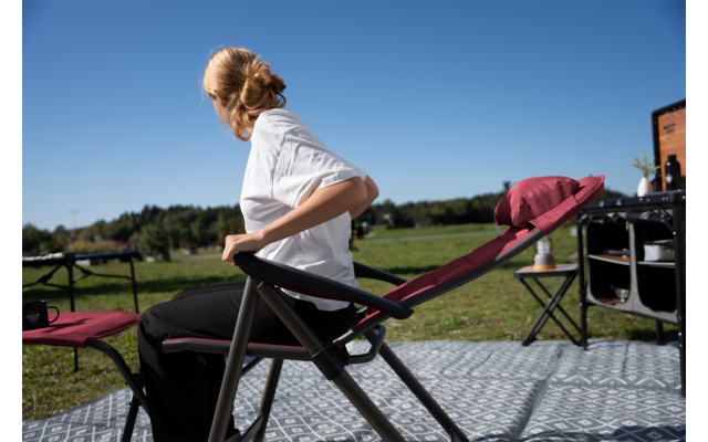 Personne aux cheveux blonds attachés, en t‑shirt blanc et pantalon noir, assise sur une chaise pliante rouge et regardant vers la gauche. Tapis à motifs au premier plan, champ vert et ciel bleu en arrière‑plan, deuxième chaise et matériel de camping près d’un bâtiment en bois
