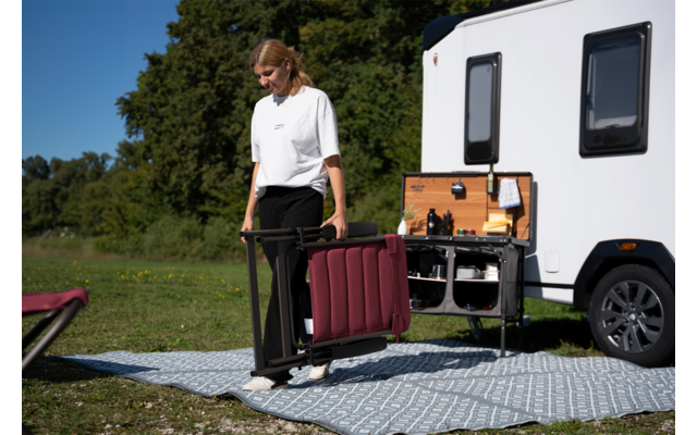 Personne portant une chaise de camping pliée bordeaux à côté d'un camping-car blanc avec une cuisine extérieure ouverte, tapis à motifs sur l'herbe, arbres et ciel bleu en arrière-plan