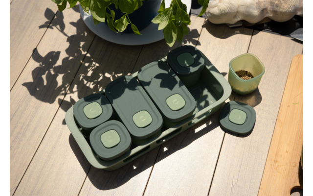 Meerdere groene bakjes met vierkante deksels in een bijpassend bakje op een houten oppervlak, linksboven een pot met groene plant, rechts een klein potje met aarde en rechtsboven een lichtgekleurde steen