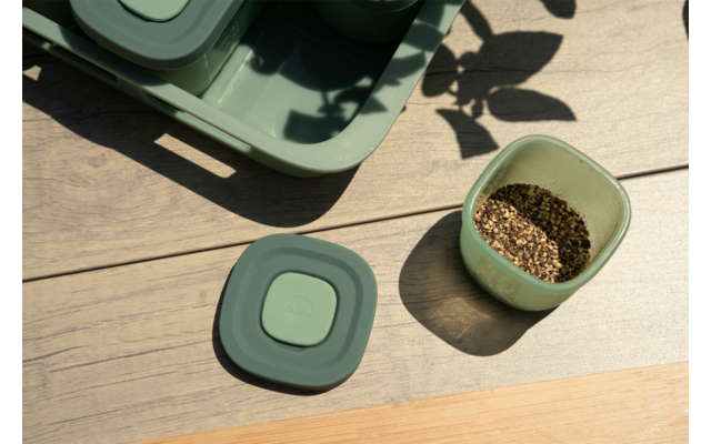 Houten oppervlak met twee groene vierkante bakjes, één open met bruine korrels, een deksel ernaast; linksboven een groen dienblad en bladschaduwen