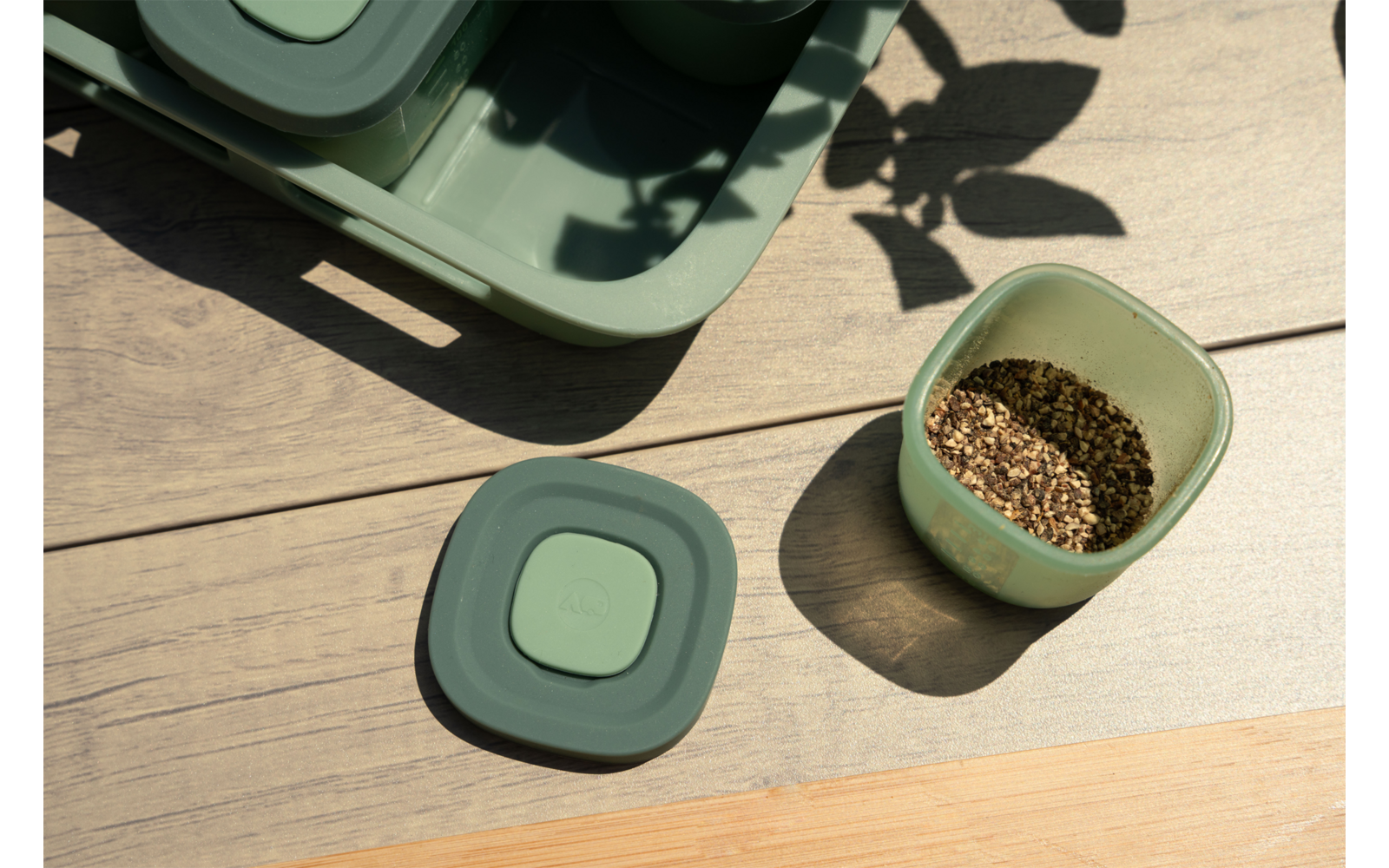 Houten oppervlak met twee groene vierkante bakjes, één open met bruine korrels, een deksel ernaast; linksboven een groen dienblad en bladschaduwen