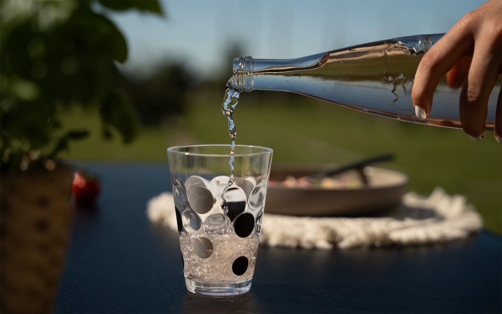 Hand pouring water from a glass bottle into a glass with black dots on an outdoor table; potted plant and plate on a placemat blurred in the background