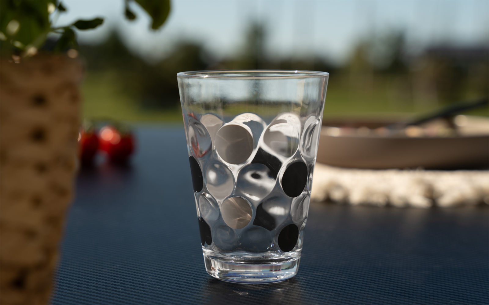Clear drinking glass with circular pattern on a black table, blurred background with a plant pot, cherry tomatoes and a bowl