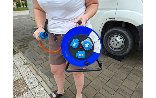 Person vom Brust- bis Oberschenkelbereich hält eine blaue Kabeltrommel mit drei Steckdosen und orangefarbenem Kabel; im Hintergrund ein weißes Fahrzeugrad auf Pflaster