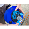 Hände halten eine blaue Kabeltrommel mit geöffneten Steckdosenabdeckungen und einem orangefarbenen Kabel auf gepflastertem Boden