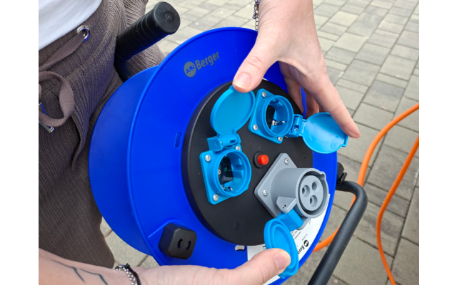 Hände halten eine blaue Kabeltrommel mit geöffneten Steckdosenabdeckungen und einem orangefarbenen Kabel auf gepflastertem Boden