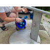 Teilansicht einer knienden Person, Hände halten eine blaue Kabeltrommel mit Steckdosen neben einem Metallwasserstandrohr mit Wasserhahn, Bodenplatten und Bäume im Hintergrund