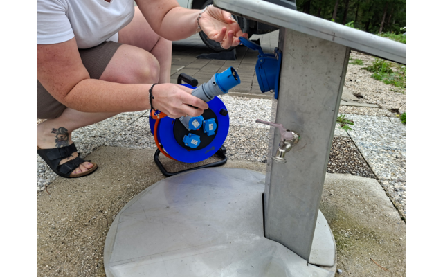 Teilansicht einer knienden Person, Hände halten eine blaue Kabeltrommel mit Steckdosen neben einem Metallwasserstandrohr mit Wasserhahn, Bodenplatten und Bäume im Hintergrund