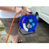Hände halten eine blaue Kabeltrommel mit orangefarbenem Kabel; Person trägt weißes Oberteil, braune Shorts und schwarze Sandalen; weißes Wohnmobil und gepflasterter Boden im Hintergrund