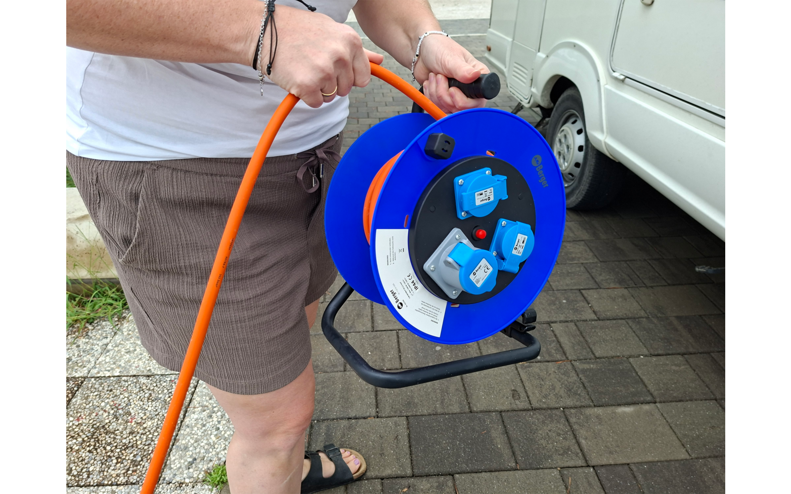 Hände halten eine blaue Kabeltrommel mit orangefarbenem Kabel; Person trägt weißes Oberteil, braune Shorts und schwarze Sandalen; weißes Wohnmobil und gepflasterter Boden im Hintergrund