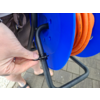 Hand hält einen schwarzen Kabelbinder um das schwarze Metallgestell einer blauen Kabeltrommel mit orangefarbenem Kabel; Pflasterboden und Beine mit Sandalen und Shorts sichtbar; Rucksackriemen am Rand